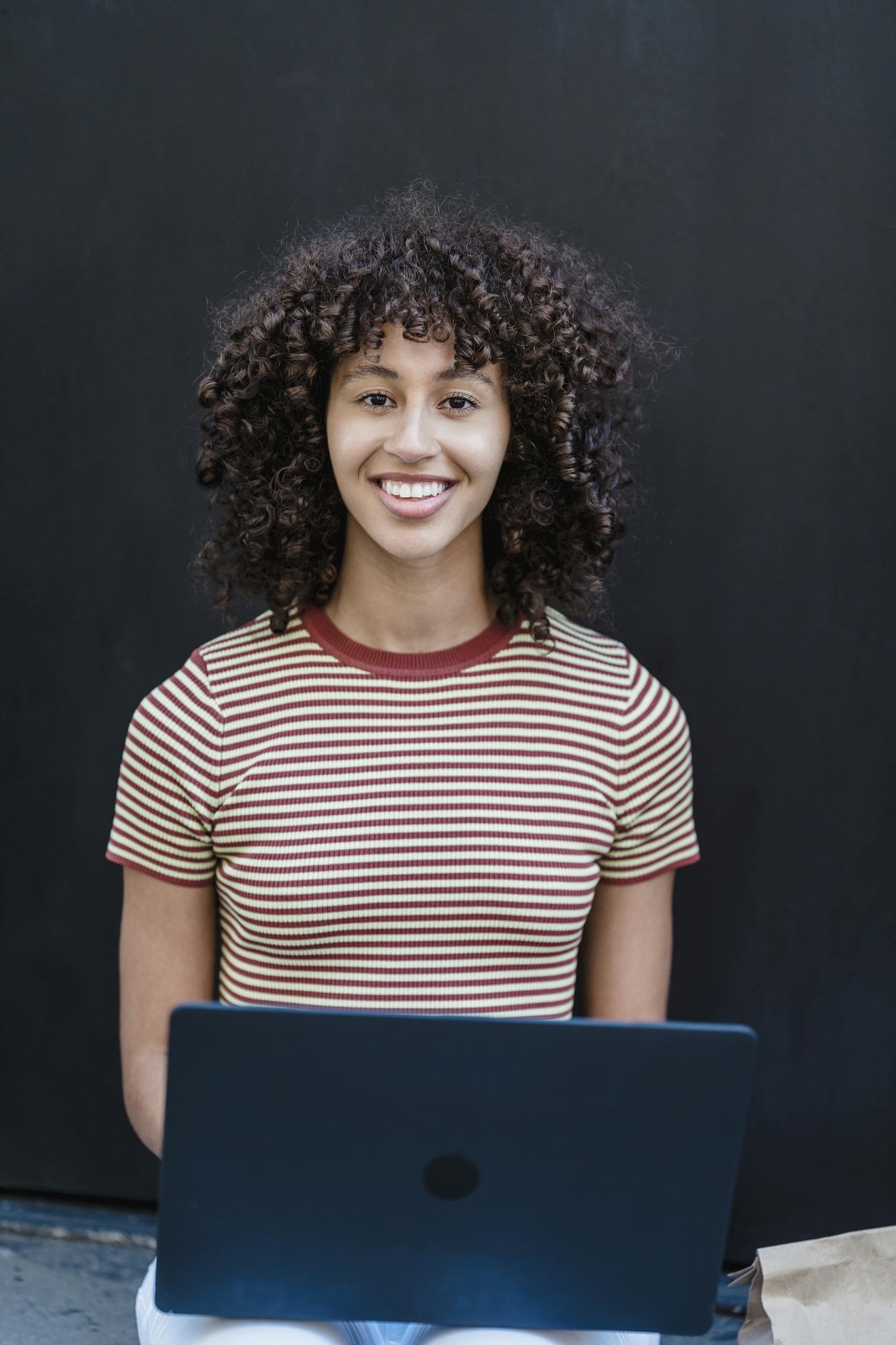 Smiling young woman with curly hair works on a laptop outdoors, embracing a casual and trendy lifestyle.